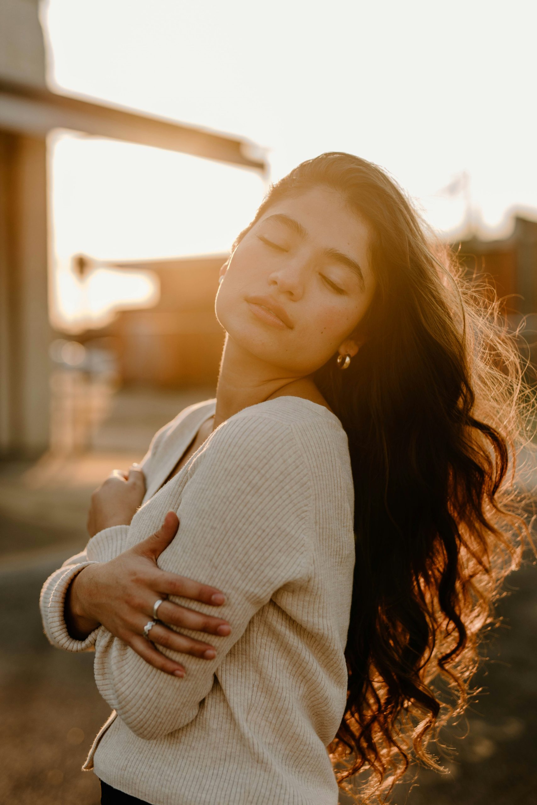A woman standing outdoors with her arms wrapped around herself, eyes closed in the evening light